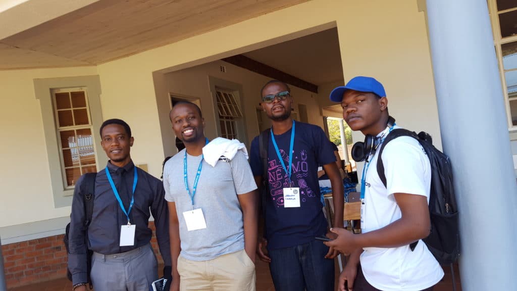 4 junge Teilnehmer posieren für ein Foto; dunkle Haut, von leger bis formell gekleidet, einer mit Base-Cap, alle mit WordCamp-Namensschildern um den Hals
